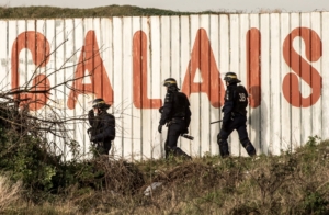 French riot police officer walk in front of a fence with the lettering 'Calais' near the A16 motorway near the site of the Eurotunnel in Coquelles, near Calais, northern France on January 21, 2016. - Approximately 300 migrants have tried to board trucks protected by French police, according to an estimate made by an AFP photographer present at the scene. Clashes already erupted briefly on the night of January 20 at the port bypass Calais between several hundred migrants and security forces, who fired tear gas to restore the situation, according to an AFP correspondent. These incidents occurred after the prefecture of Pas-de-Calais had set an ultimatum which expired early in the afternoon for the last migrants to leave a deforested 100 metre strip of the "Jungle" camp along the ring road for safety reasons. (Photo by PHILIPPE HUGUEN / AFP)
