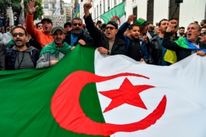 People chant slogans as they wave a large Algerian national flag during a weekly anti-government demonstration in the capital Algiers on March 6, 2020. - Protests against cronyism have continued in Algeria despite former president Abdelaziz Bouteflika's resignation and the election of a new president in December 2019.